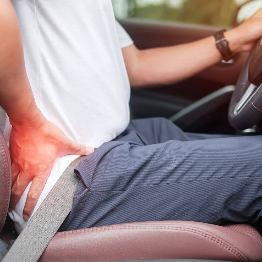 A person in a car is holding their lower back, appearing uncomfortable, with a seatbelt fastened.