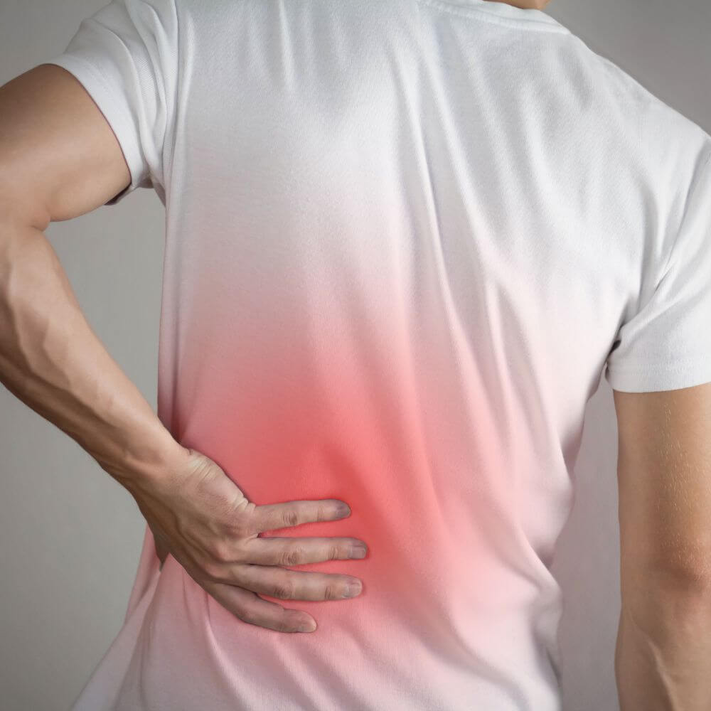 A person wearing a white shirt is holding their lower back, indicating discomfort or pain, with a red gradient highlighting the affected area.