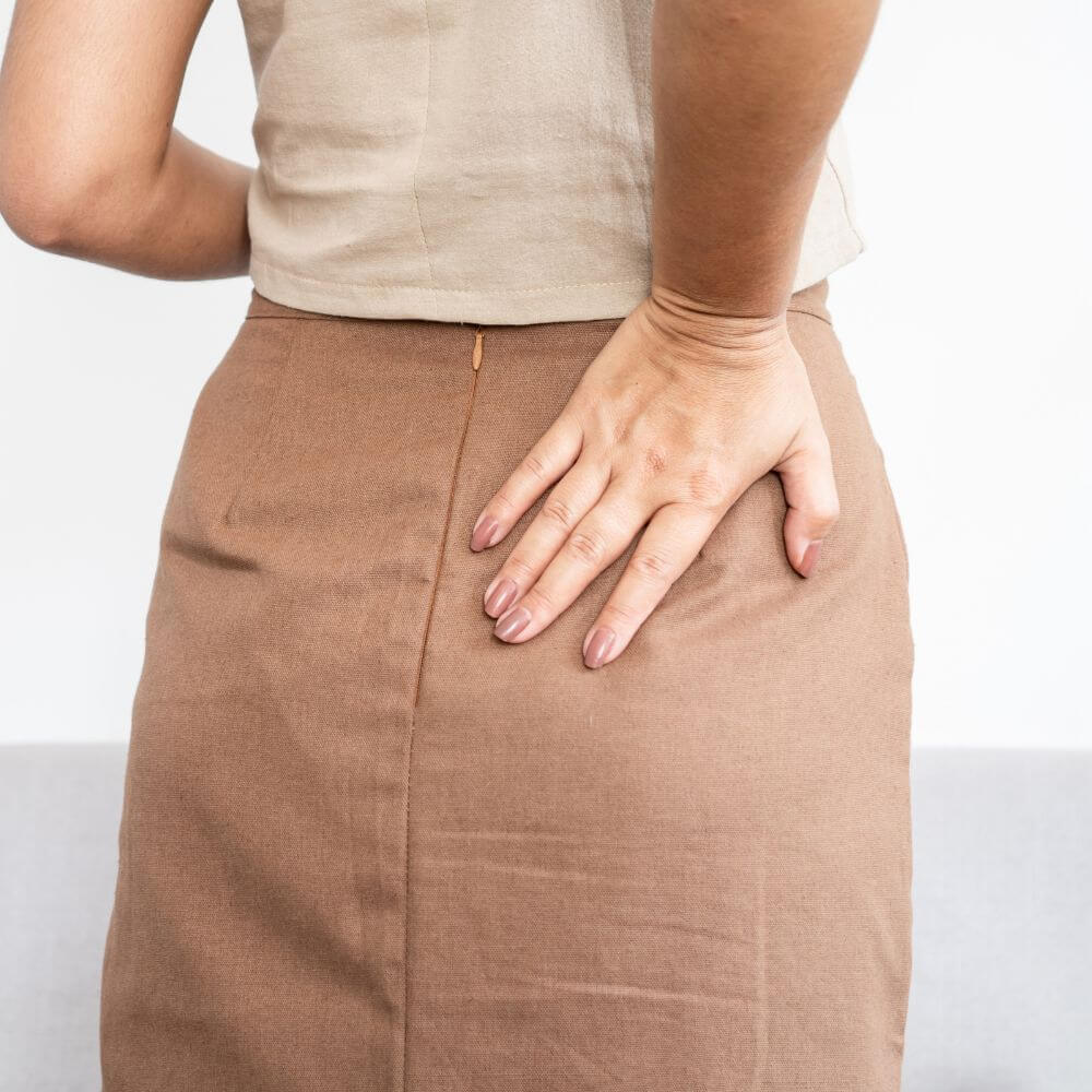 A person with their back to the camera is wearing a beige top and a brown skirt, with one hand resting on their hip.